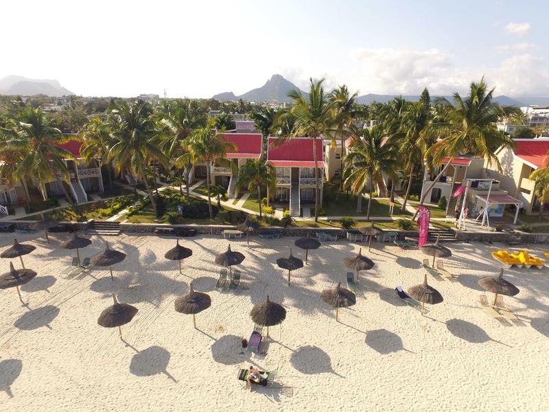 Pláž s mnoha slunečníky a palmami před bungalovy za slunečného dne.