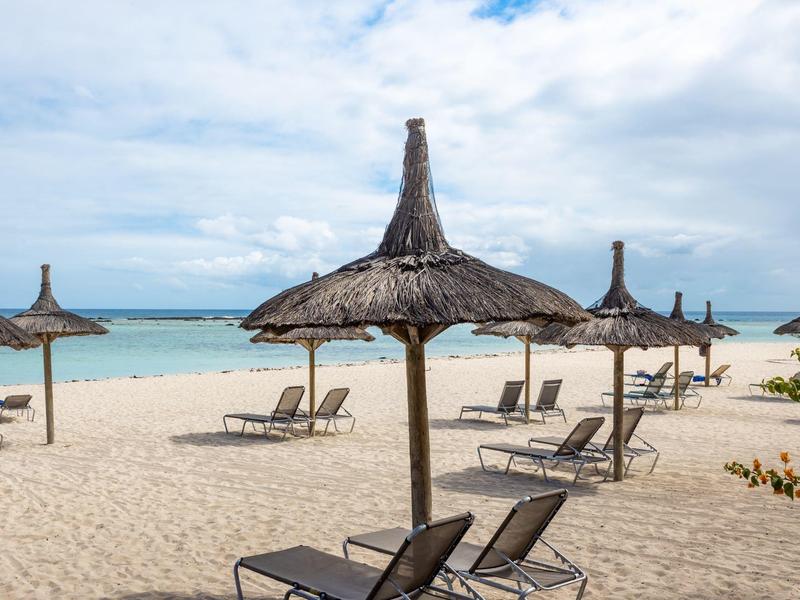 Pláž s pískem, slámovými slunečníky a lehátky u klidné tyrkysové vody.