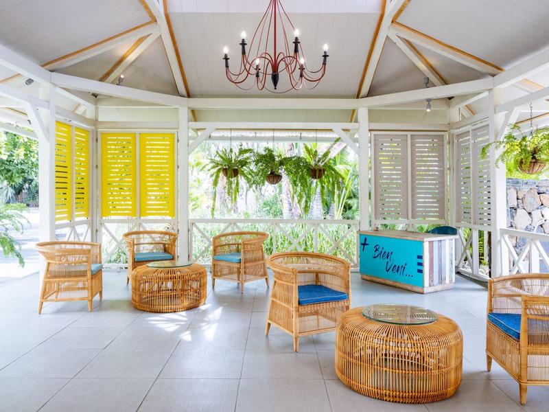 Světlá lounge s ratanovým nábytkem, modrými polštáři a závěsnými rostlinami.