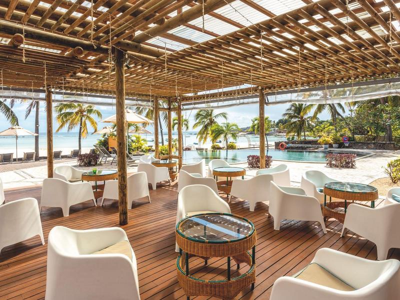 Otevřená restaurace s dřevěnou podlahou a bílými židlemi, výhled na palmy a pláž.