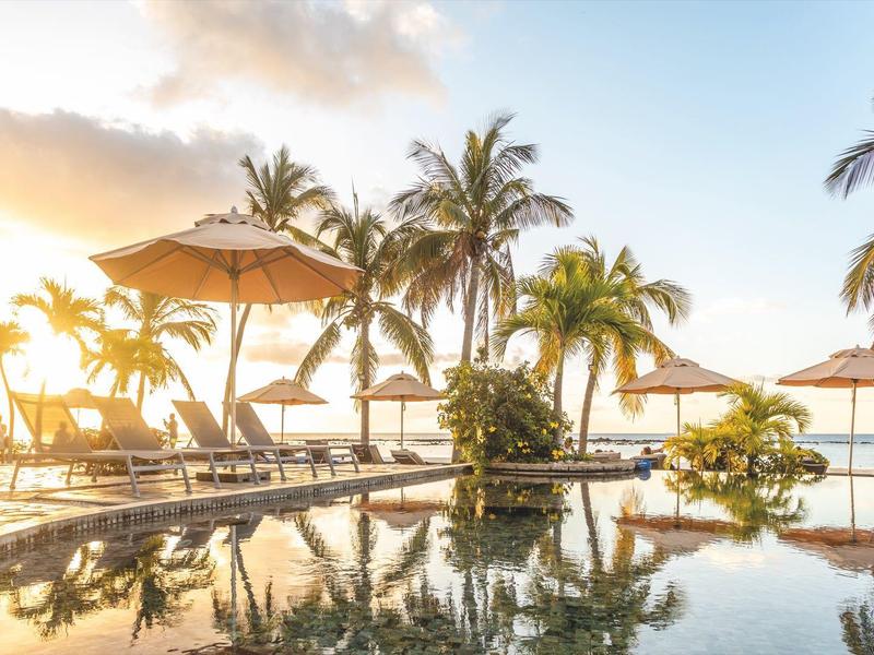 Západ slunce nad rezortním bazénem s palmami a lehátky na pláži.