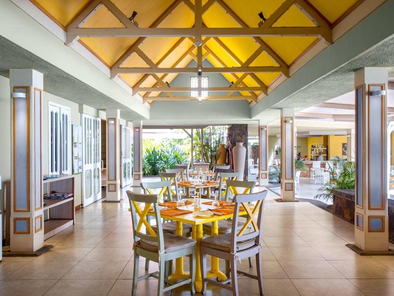 Prosvětlená otevřená restaurace s béžovou podlahou, dřevěným stropem a stoly se žlutými židlemi.