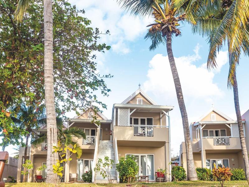 Dům s verandou obklopený palmami a zelenými rostlinami pod modrou oblohou.