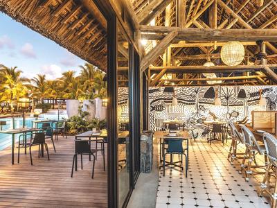 Offenes Restaurant mit Holzdach, weißen Fliesenboden, modernen Tischen und Blick auf Pool und Palmen.