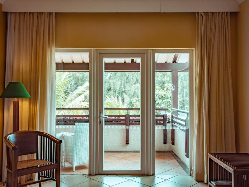 Výhled na balkón přes skleněné dveře v útulném hotelovém pokoji se židlí a stojací lampou.