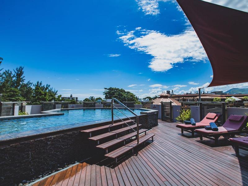 Moderní bazén s lehátky a sluneční plachtou na dřevěné terase za jasné oblohy.