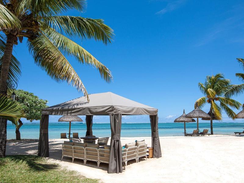 Strand mit weißen Sand, Palmen und einer überdachten Lounge mit Sitzgelegenheiten unter blauem Himmel.