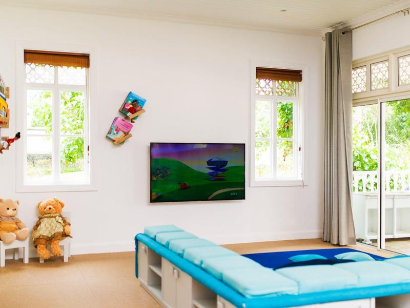 Helles Wohnzimmer mit blauem Sofa, TV an der Wand, Kuscheltieren und großen Fenstern.