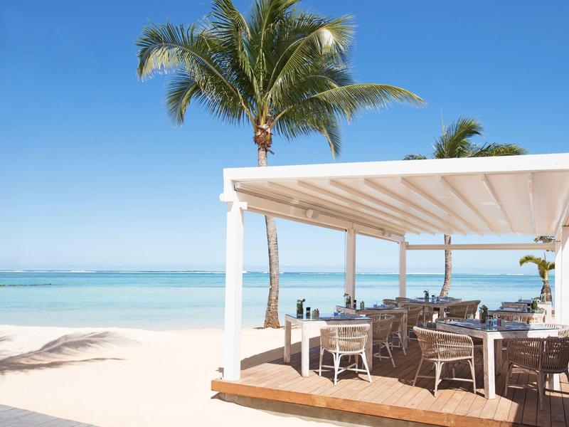 Strand mit weißem Sand, klarem blauen Meer, Palmen und einem offenen Restaurant mit Holzstühlen.