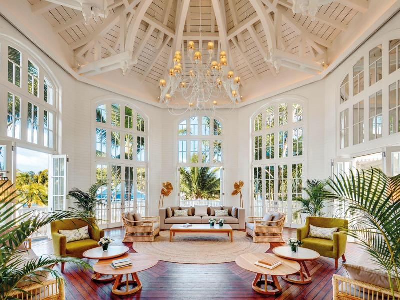 Heller Wintergarten mit hohen weißen Fenstern, Holzbalkendecke, Pflanzen und gemütlichen Sitzmöbeln.