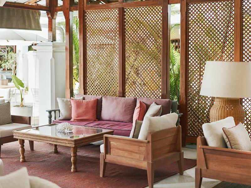 Světlý, útulný hotelový obývací pokoj se dřevěným nábytkem, růžovými polštáři a stojací lampou.