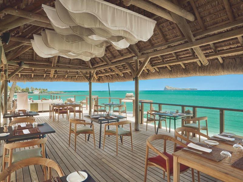 Otevřená restaurace s dřevěným nábytkem pod střechou z palmových listů s výhledem na tyrkysové moře.