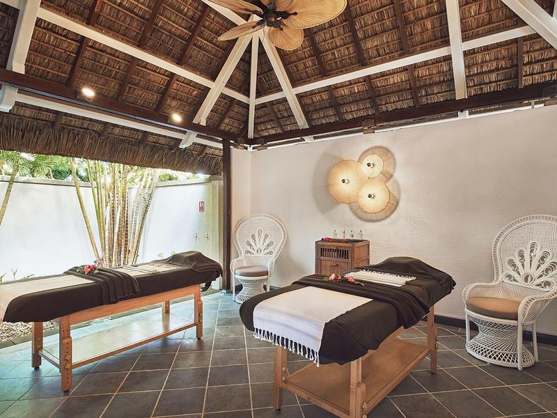 Světelná spa místnost s masážními stoly, židlemi a stropním ventilátorem pod doškovou střechou.