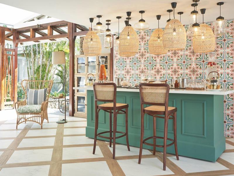 Světlý hotelový bar se zeleným pultem, dvěma dřevěnými stoličkami, ratanovými lampami a vzorovanou stěnou.