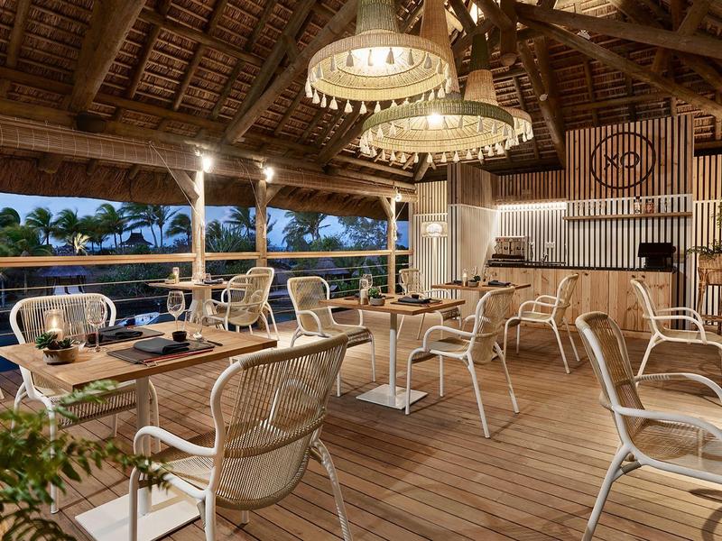 Otevřená restaurace s dřevěným stropem, několika stoly a židlemi a tropickým výhledem.