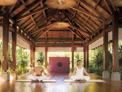 Zwei Personen meditieren auf Holzboden in offenem Raum mit Holzdach und Pflanzen im Hintergrund.