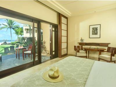 Helles Schlafzimmer mit großem Glasfenster, Blick auf Meer, Terrasse, Holzmöbel und Strohhut auf Bett.