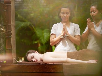 Frau liegt entspannt auf Massageliege, während Therapeutin Hände in Gebetshaltung hält im grünen Umfeld.