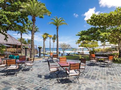 Offener Sitzbereich mit Stühlen und Tischen unter Palmen auf gepflastertem Boden am Meer.