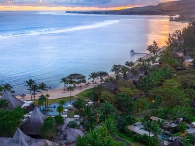 Strand mit Palmen, grüner Vegetation und mehreren Hütten am Ufer bei Sonnenuntergang.