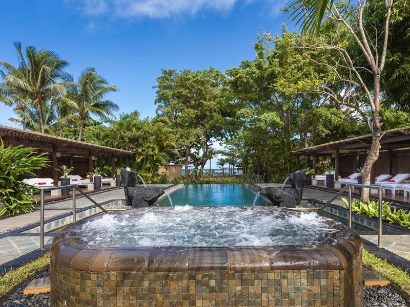 Außenjacuzzi mit sprudelndem Wasser vor Pool, umgeben von Palmen und gemütlichen Liegen.