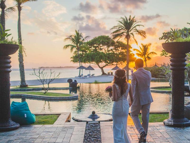 Paar in weißen Kleidern steht vor Pool mit Palmen und Sonnenuntergang am Meer.