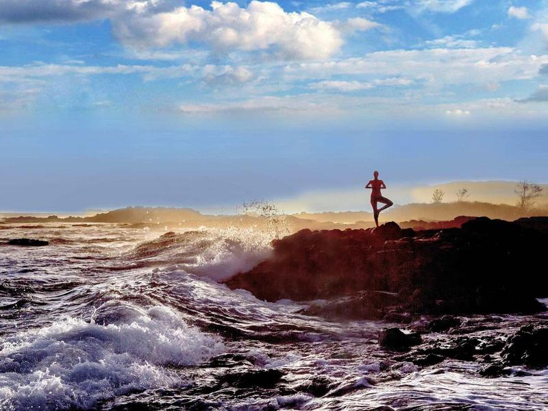 Eine Person steht auf felsiger Küste, umgeben von wildem Meer und blauem Himmel mit Wolken.