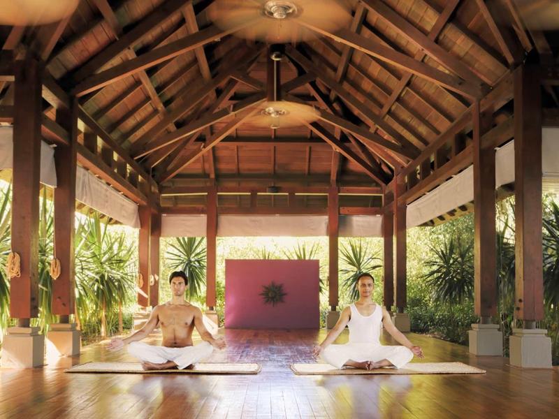 Zwei Personen meditieren auf Holzboden in offenem Raum mit Holzdach und Pflanzen im Hintergrund.