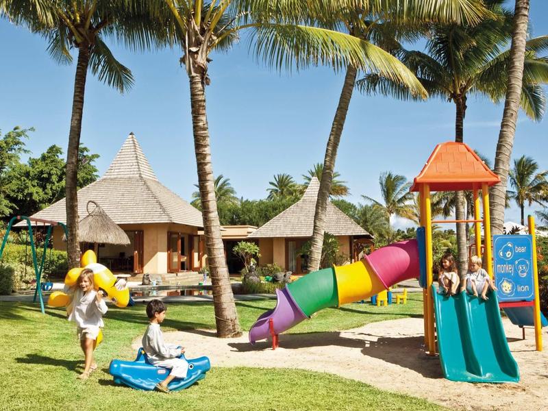 Kinder spielen auf einem bunten Spielplatz mit Rutsche und Schaukel unter Palmen.