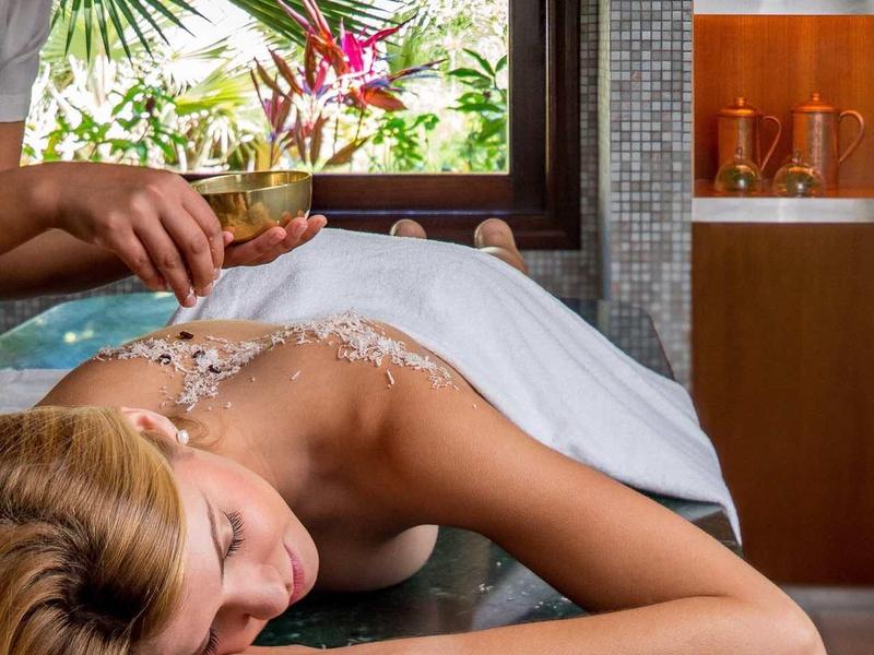 Frau liegt entspannt auf Massagetisch, Massagetherapeutin trägt Öl auf Rücken auf, helle, natürliche Umgebung.