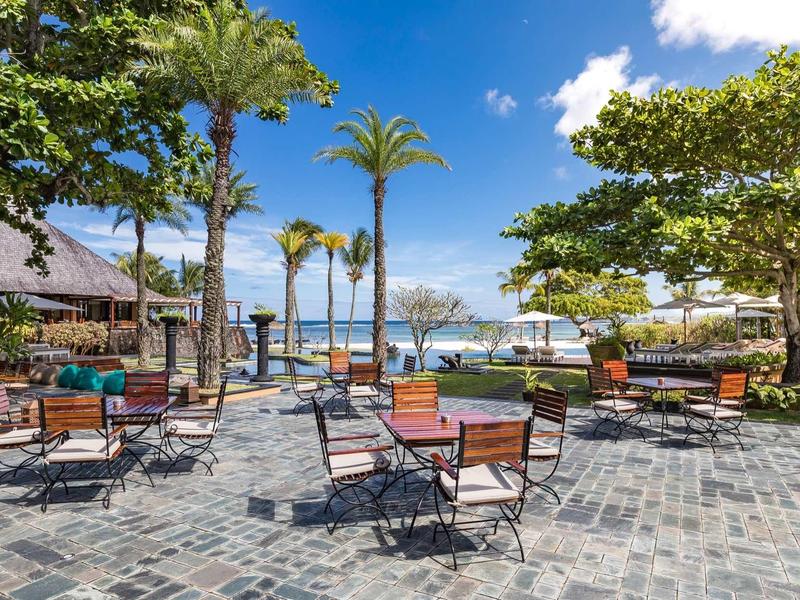 Offener Sitzbereich mit Stühlen und Tischen unter Palmen auf gepflastertem Boden am Meer.