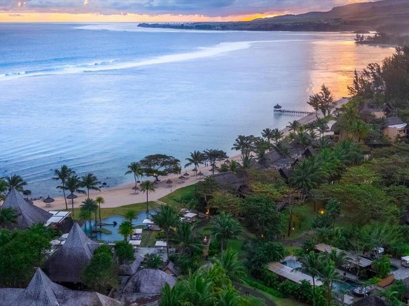 Strand mit Palmen, grüner Vegetation und mehreren Hütten am Ufer bei Sonnenuntergang.