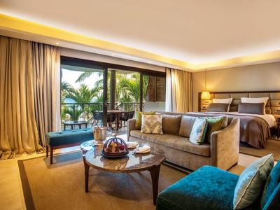 Habitación de hotel lujosa con área de descanso, cama grande y vista a una terraza con palmeras.