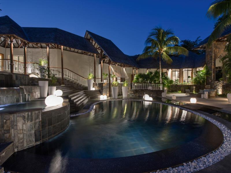 Hotel lujoso con piscina iluminada y jardín tropical por la noche.