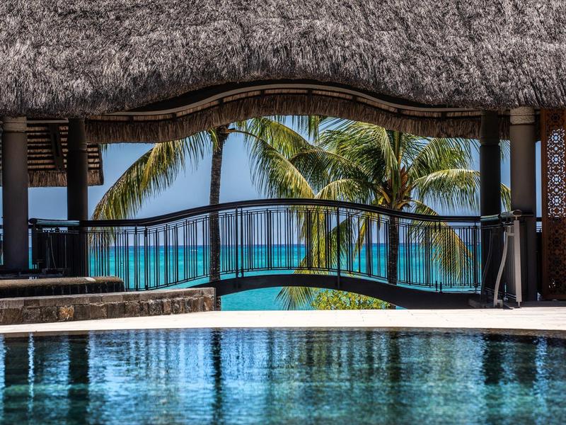 Vista de una piscina con un bar techado, palmeras y el mar al fondo.