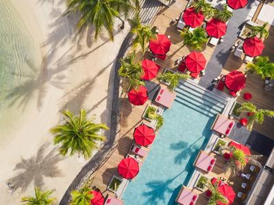 Luftaufnahme eines Pools mit roten Sonnenschirmen neben einem Sandstrand und Palmen.