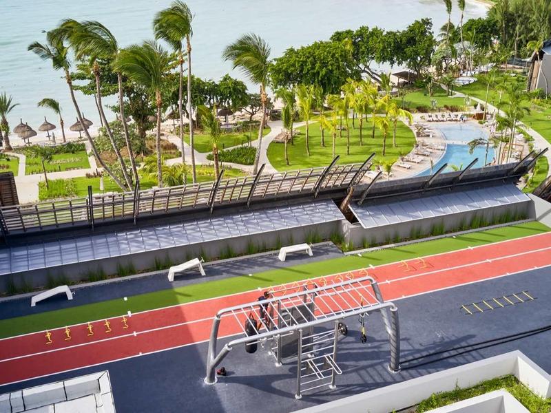 Blick auf ein gepflegtes Sportgelände mit Laufbahn, Fitnessgeräten, Palmen und Strand im Hintergrund.