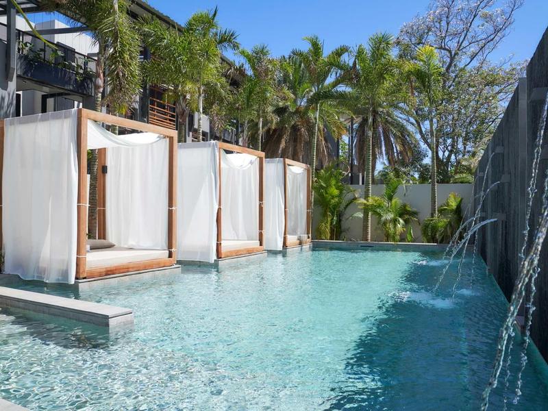Moderner Pool mit Wasserspielen, weißen Liegebetten und Palmen im Hintergrund bei sonnigem Wetter.