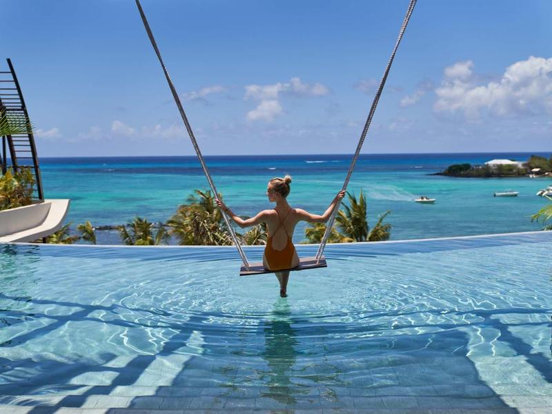 Frau schaukelt über Infinity-Pool mit Blick auf türkisfarbenes Meer und blauen Himmel.