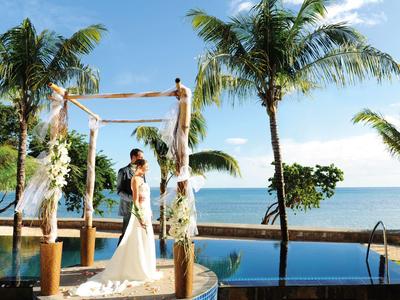 Bruidegom en bruid onder een versierde boog met palmbomen en zee op de achtergrond