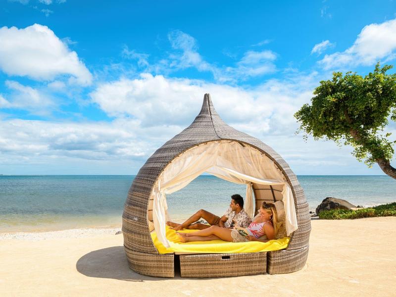 Pár odpočívá v útulném kulatém plážovém lehátku na slunečné pláži s výhledem na moře.