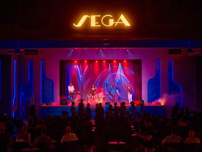 Palco illuminato con musicisti e pubblico davanti all'hotel SEGA di notte.