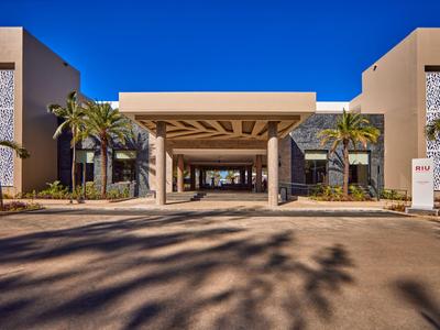 Ingresso moderno dell'hotel con ampio portico, palme e cielo azzurro limpido.