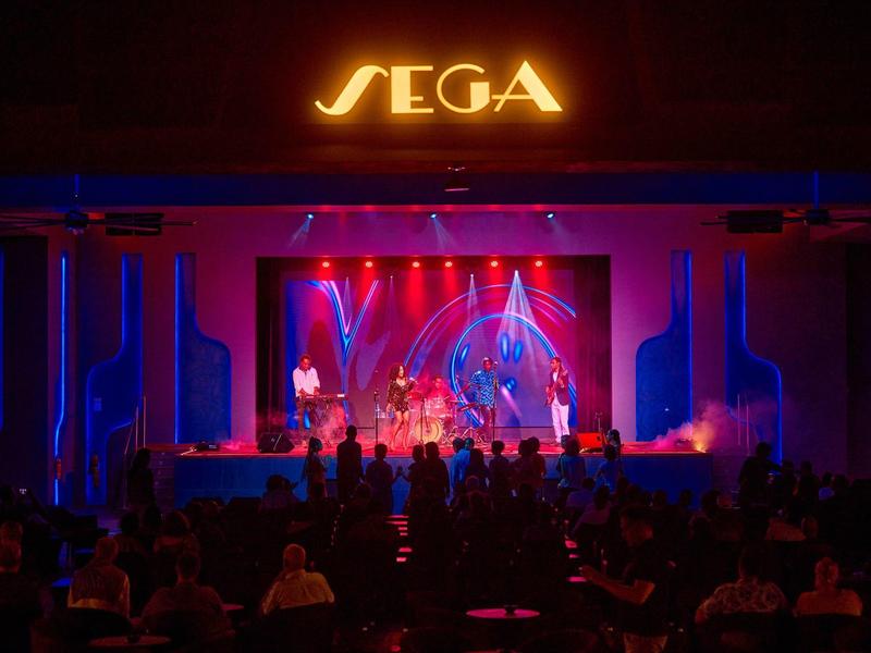 Bühne mit Beleuchtung und SEGA-Logo, Publikum vor der Bühne bei einer Veranstaltung.