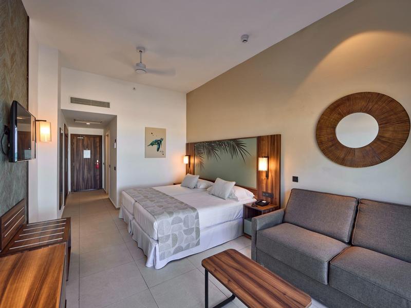 Modern eingerichtetes Hotelzimmer mit Doppelbett, Sofa und Holzmöbeln in warmen Farbtönen.