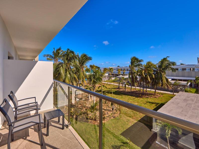 Balkon mit Stühlen und Glasgeländer, Blick auf palmengesäumten Garten und weiße Gebäude