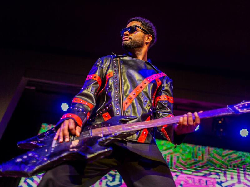 Mann mit Sonnenbrille spielt auf einer Bühne E-Gitarre unter bunten Lichtern.