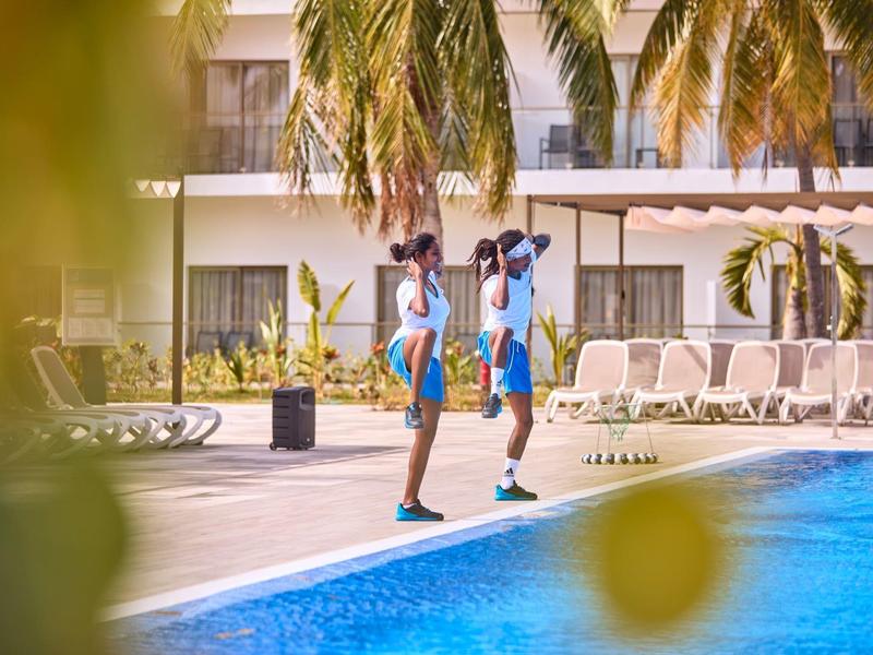 Zwei junge Frauen in Sportkleidung gehen am Pool eines Hotels, umgeben von Liegestühlen und Palmen.