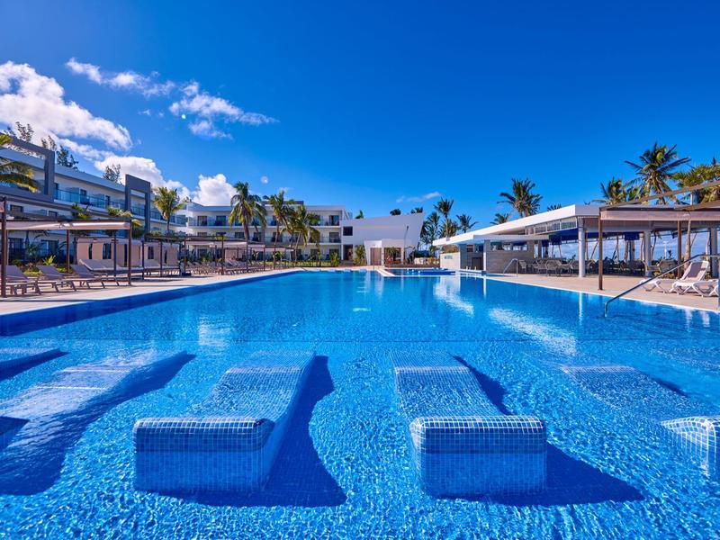 Großer Hotelpool mit eingebauten Liegen, umgeben von Palmen und Sonnenschirmen unter klarem Himmel.