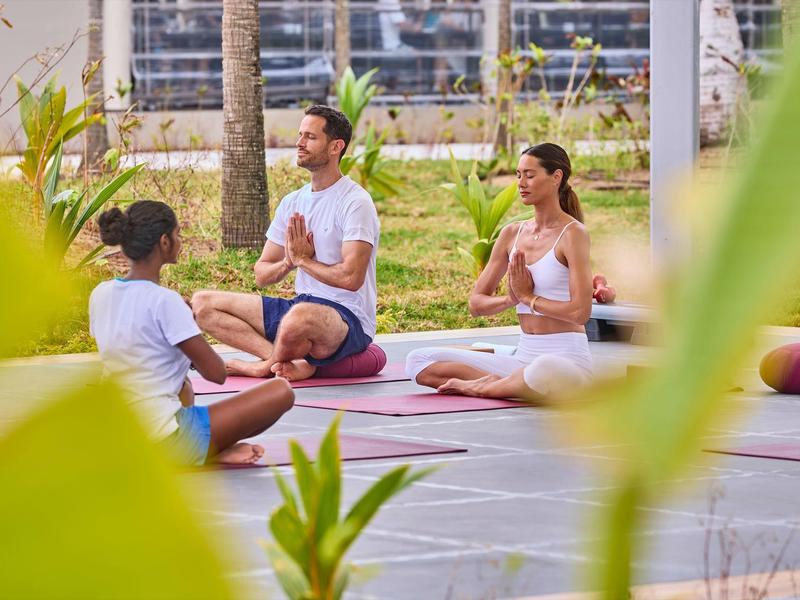 Drei Personen praktizieren Yoga im Freien, sitzend auf Yogamatten in einer entspannten Haltung.